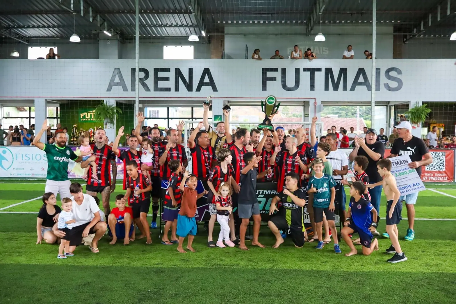 Time comemorando com o troféu ao lado de crianças e familiares na quadra